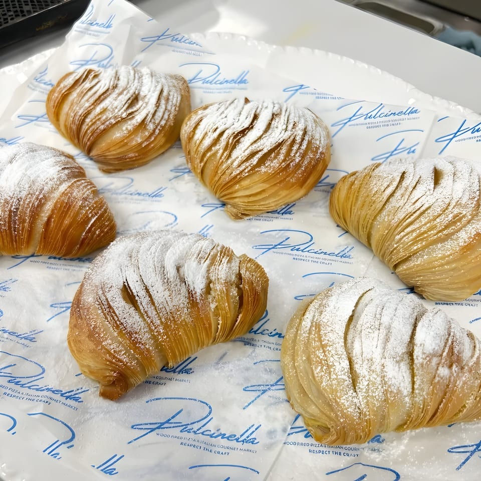 Six powdered sugar-dusted sfogliatelle pastries on white paper with blue Pulcinella branding.