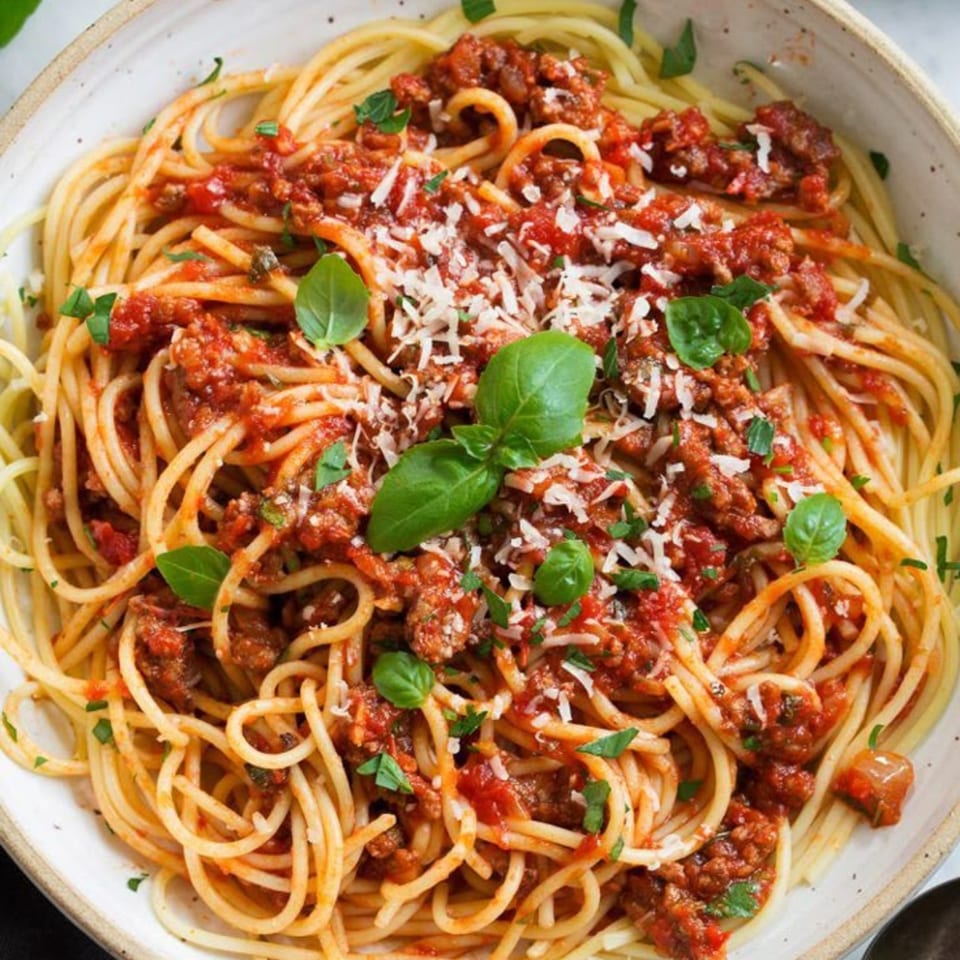 Close-up of spaghetti with rich red meat sauce, grated Parmesan, and fresh basil in a white bowl.