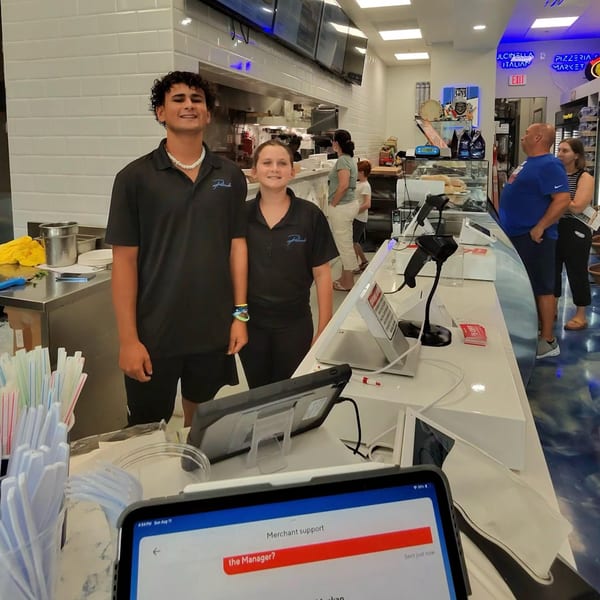 Two Pulcinella Pizza employees stand behind a counter with customers and kitchen in the background.