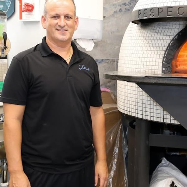 Man in black polo shirt smiling next to a white tiled pizza oven with glowing embers.