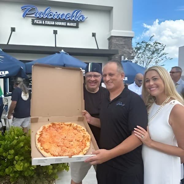 Three people smiling, holding a large cheese pizza in a box outside Pulcinella Pizza and Italian Market.