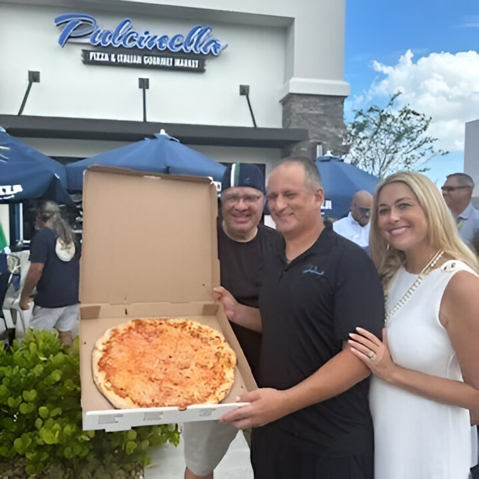 Three people smiling, holding a large cheese pizza in a box outside Pulcinella Pizza and Italian Market.