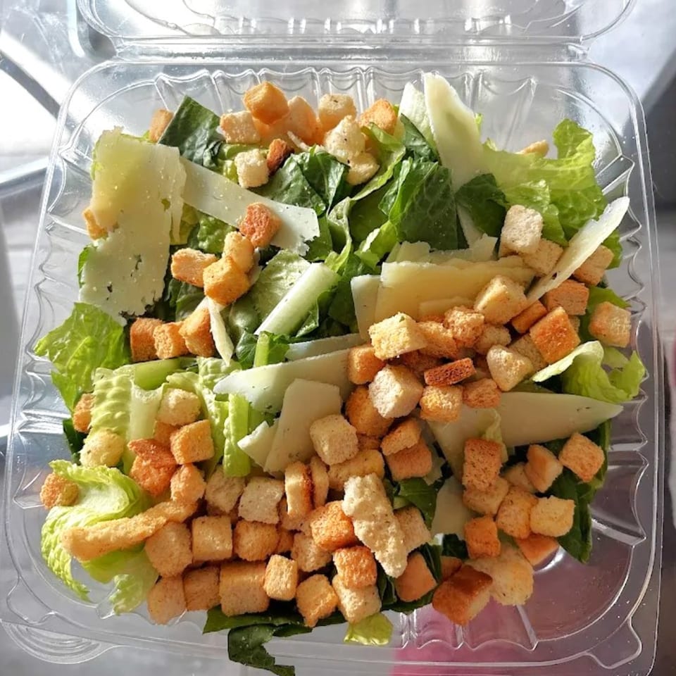 Caesar salad with romaine lettuce, shaved Parmesan, and croutons in a clear plastic container.