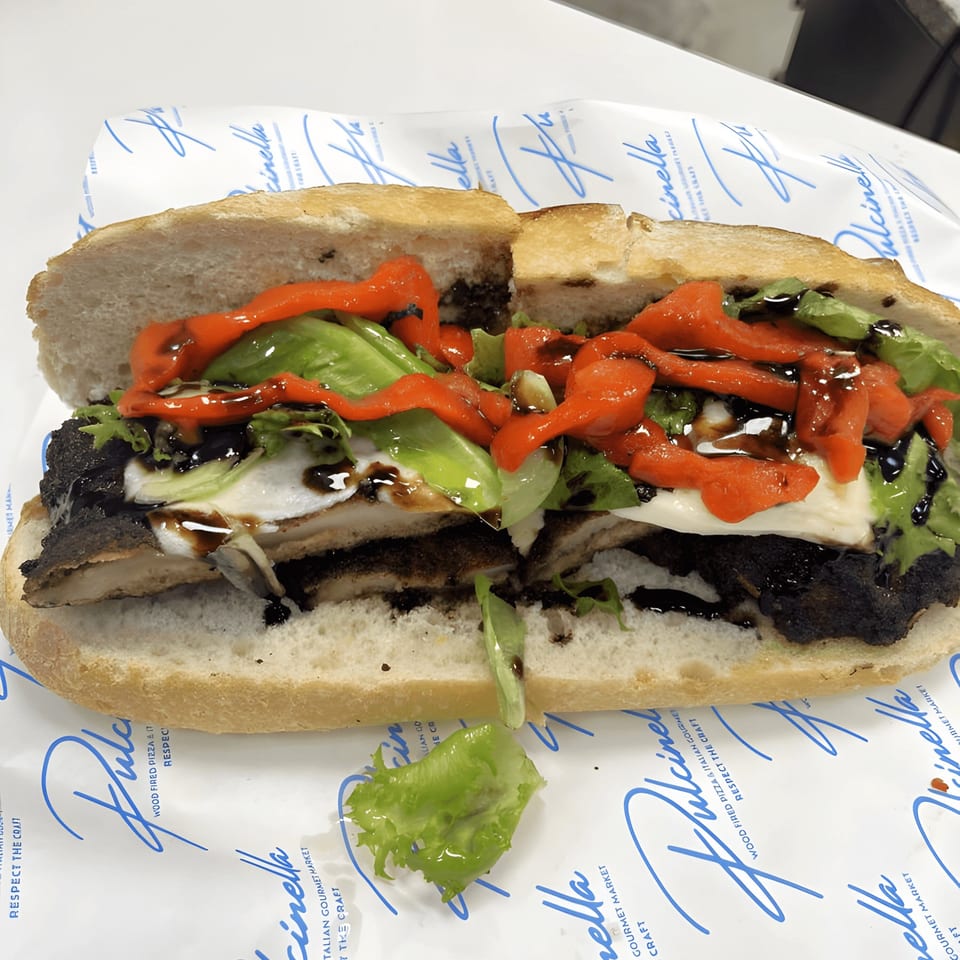 A Pulcinella Italian sub sandwich with red peppers, lettuce, and balsamic glaze on branded paper.