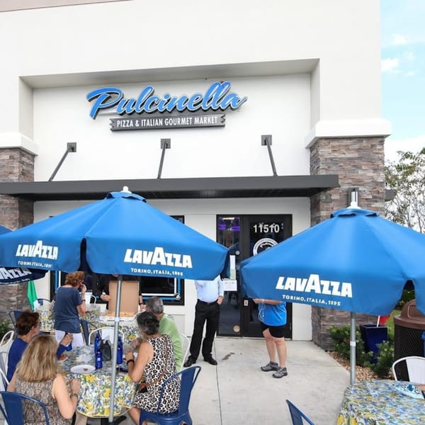 Pulcinella Pizza & Italian Market storefront with people dining outdoors under blue Lavazza umbrellas.