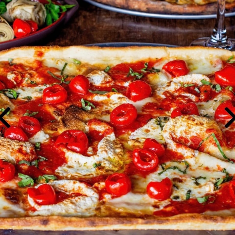 Rectangular pizza with red sauce, white cheese, small red peppers, and basil on a wooden table.