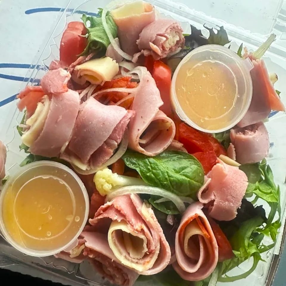 A deli salad with rolled meats and cheeses, lettuce, tomatoes, onions, and two dressing cups.